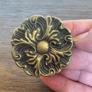 May include: Ornate, antique-style brass cabinet knob with a floral design. The knob features a central circular element surrounded by detailed, swirling leaf patterns. The finish has a slightly aged appearance, with darker accents in the recesses.