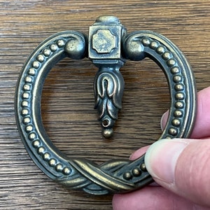 May include: A decorative, ornate, brass-toned, ring-shaped pull with a beaded edge and a floral design.