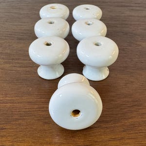 May include: Eight white ceramic cabinet knobs with a glossy finish are arranged on a wooden surface. Each knob has a rounded top with a small hole and a pedestal base. The knobs are a classic design for furniture.