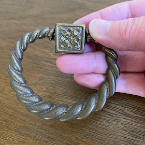 May include: Antique brass-toned drawer pull with a twisted rope design. The square center piece features a floral pattern. The pull is circular and appears to be made of metal. The hardware is designed for furniture.