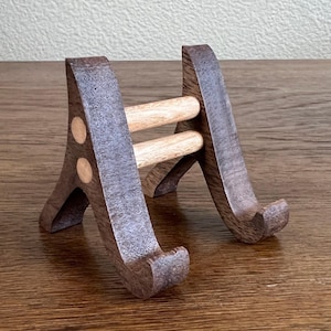 May include: A wooden phone stand with two light brown wooden supports and a dark brown wooden base. The stand is designed to hold a phone upright.
