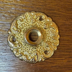 May include: Ornate gold-colored door knob backplate with a central hole and three smaller holes. The backplate features a detailed floral and scroll design, adding a vintage aesthetic. The metal has a polished finish.