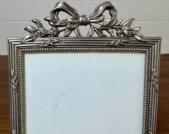 May include: A silver-toned rectangular picture frame with a bow and floral design at the top. The frame has a beaded border and a white interior, suitable for a photograph. The frame is made of metal and has a vintage aesthetic.