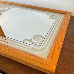 May include: Rectangular mirror with a light brown wooden frame. The mirror's surface features a decorative design with geometric shapes and scrollwork. The mirror is likely for wall decor.