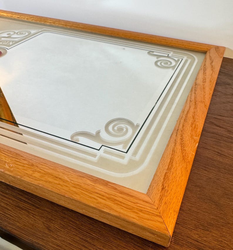 May include: Rectangular mirror with a light brown wooden frame. The mirror's surface features a decorative design with geometric shapes and scrollwork. The mirror is likely for wall decor.