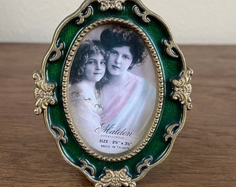 May include: A gold-colored oval picture frame with a green enamel finish. The frame has ornate details and a beaded border. It features a black and white photo of two women. The frame is labeled "Halden International" and "Size: 2 1/2" x 3 1/2" Made in Taiwan."