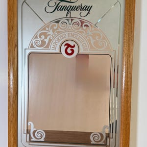May include: A framed mirror with the Tanqueray logo and decorative accents. The mirror features the words "IMPORTED ENGLISH GIN" and a red "2" symbol. The frame is made of light brown wood.