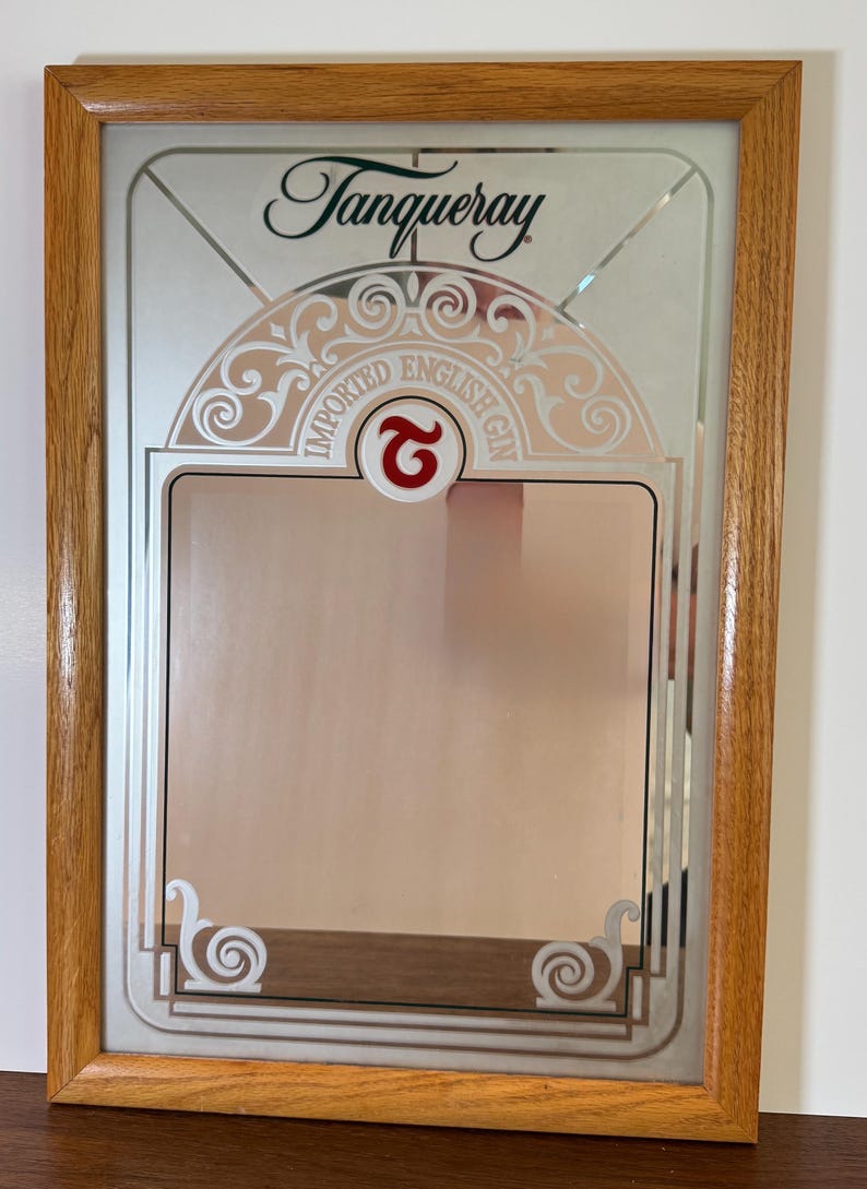 May include: A framed mirror with the Tanqueray logo and decorative accents. The mirror features the words "IMPORTED ENGLISH GIN" and a red "2" symbol. The frame is made of light brown wood.