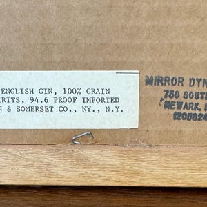 May include: Close-up of the back of a framed artwork. A white label with black text reads "IMPORTED ENGLISH GIN, 100% GRAIN NEUTRAL SPIRITS, 94.6 PROOF IMPORTED." The frame is made of light-colored wood.