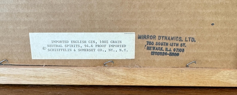 May include: Close-up of the back of a framed artwork. A white label with black text reads "IMPORTED ENGLISH GIN, 100% GRAIN NEUTRAL SPIRITS, 94.6 PROOF IMPORTED." The frame is made of light-colored wood.