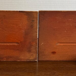 May include: A pair of wooden bookends with a simple design. Each bookend has a rectangular base with a small, rounded foot at the bottom.