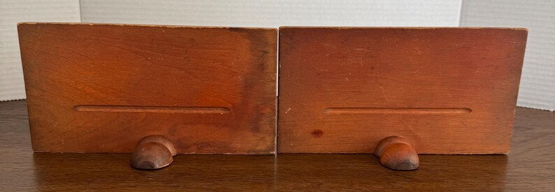 May include: A pair of wooden bookends with a simple design. Each bookend has a rectangular base with a small, rounded foot at the bottom.