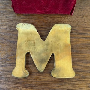 May include: A large, golden-colored metal letter "M" rests on a wooden surface. The letter has a classic serif font with rounded edges. A red velvet pouch is visible in the background.