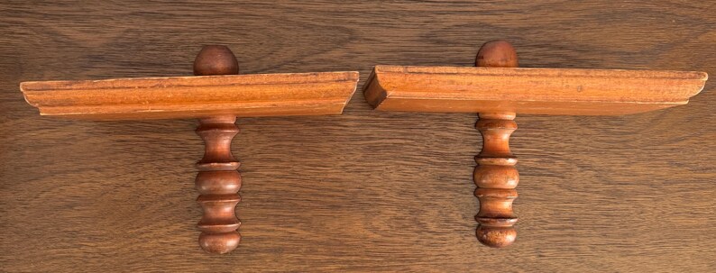 May include: Two wooden shelf brackets with a decorative, turned, spindle design. The brackets are made of light brown wood and have a simple, traditional style.