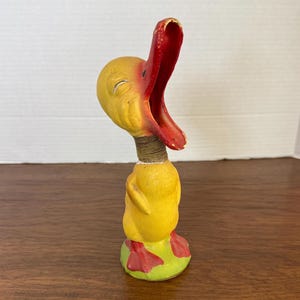 May include: A vintage yellow duck figurine with a red beak and feet. The duck has a wide open beak and is standing on a green base. The duck's neck is wrapped with a brown material.