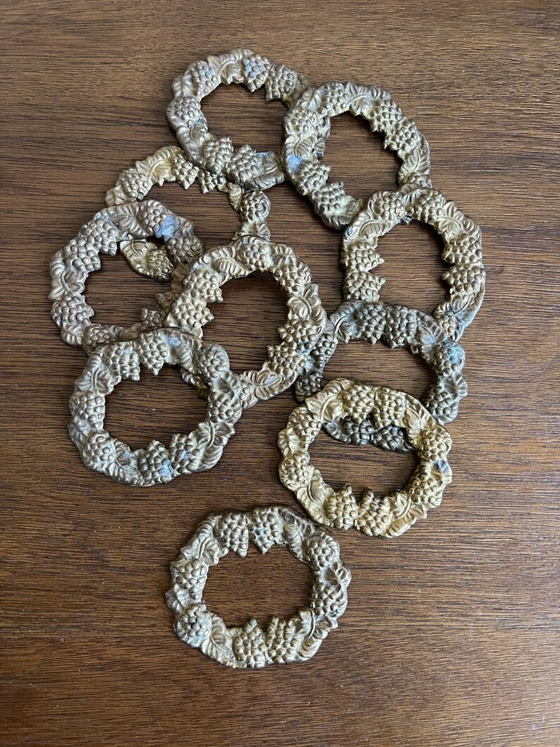 K&ouml;nnte beinhalten: Ein Set aus zehn goldfarbenen Serviettenringen mit einem Traubenmuster. Die Ringe sind verziert und detailliert, mit einer runden Form und einem erhabenen Design.