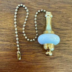 May include: A brass-colored ceiling fan pull chain with a light blue, oval-shaped accent. The chain is made of small, round beads. The pull chain is on a wooden surface.