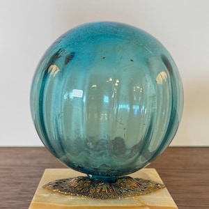 May include: A teal-colored glass sphere with vertical ridges, mounted on a decorative brass base and a rectangular marble block. The sphere has a slightly aged appearance. The base has an ornate, openwork design.