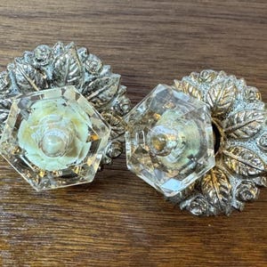 May include: Two vintage cabinet knobs. Each knob features a clear, faceted crystal center with a floral design and a silver-toned metal backing with a leaf and rose pattern. The knobs are set against a wooden surface.