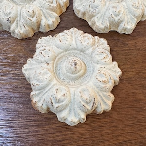 May include: Three off-white, floral-shaped decorative accents with a distressed finish. The accents are arranged on a dark wood surface. Each accent has a central circular design surrounded by petal-like shapes.