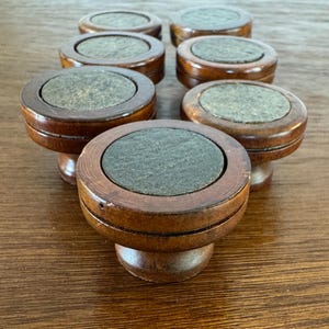 May include: A collection of seven wooden cabinet knobs with a dark brown finish. Each knob features a circular, gray inset and a rounded base. The knobs are arranged on a wooden surface, showcasing their design and craftsmanship.