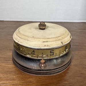 May include: Vintage metal measuring tape dispenser with a white top and black base. The tape is marked with numbers 3, 4, 5, and 6.