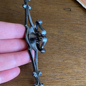 May include: Black metal drawer pull with a decorative fleur-de-lis design on each end and a ring-shaped handle in the center.