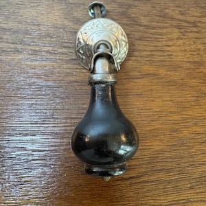 May include: A black, teardrop-shaped cabinet pull with a decorative silver-colored backplate. The backplate has an ornate, embossed design. The pull is made of a dark, glossy material and is attached to a screw.