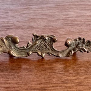 May include: Ornate antique brass drawer pull with a scrolling leaf design. The hardware is a warm, aged brass color and is set against a wooden surface. The decorative element is approximately 6 inches long.