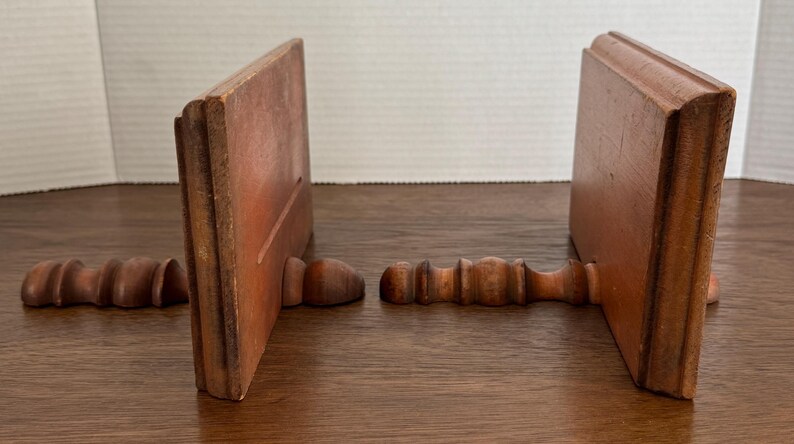 May include: Two wooden bookends with a simple design. Each bookend has a decorative, turned wooden base.