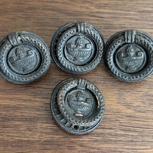 May include: Four black metal drawer pulls with a decorative design. Each pull features a circular ring with a raised design of a stylized urn with a floral motif.