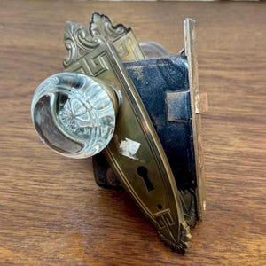 May include: An antique door lock set with a clear glass doorknob and ornate brass hardware. The set includes a keyhole and decorative detailing, showcasing a vintage aesthetic. The lock mechanism is black, and the brass has a weathered appearance.