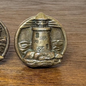 May include: Bronze-colored, circular cabinet knobs featuring a detailed relief of a lighthouse on a rocky outcropping, surrounded by stylized waves. The lighthouse has a small, square window and a domed top. The knobs are likely made of metal.