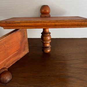 May include: A wooden shelf with a decorative, turned wooden support. The shelf is stained a reddish-brown colour and has a simple, rectangular shape.