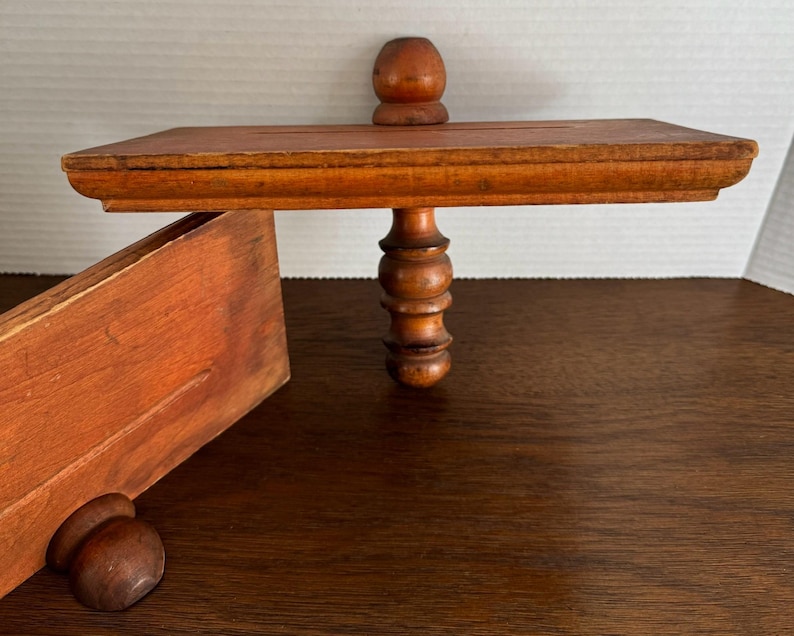 May include: A wooden shelf with a decorative, turned wooden support. The shelf is stained a reddish-brown colour and has a simple, rectangular shape.