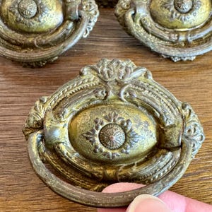 May include: Antique brass drawer pulls with ornate detailing. The pulls have a circular design with a central medallion and a curved handle. The brass has a weathered, aged appearance, with hints of green and brown. The pulls are likely for furniture restoration.
