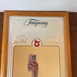 May include: Framed Tanqueray gin advertisement with a wooden frame. The advertisement features the Tanqueray logo and the words "IMPORTED ENGLISH GIN". A person is holding a pink phone in front of the advertisement.