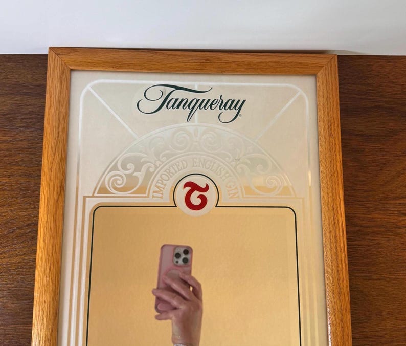 May include: Framed Tanqueray gin advertisement with a wooden frame. The advertisement features the Tanqueray logo and the words "IMPORTED ENGLISH GIN". A person is holding a pink phone in front of the advertisement.