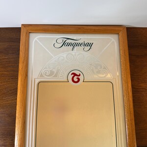 May include: A framed Tanqueray gin advertisement. The wooden frame surrounds a cream-colored background with the Tanqueray logo in elegant script. The bottom section is a gold color. The words "IMPORTED ENGLISH GIN" are visible.