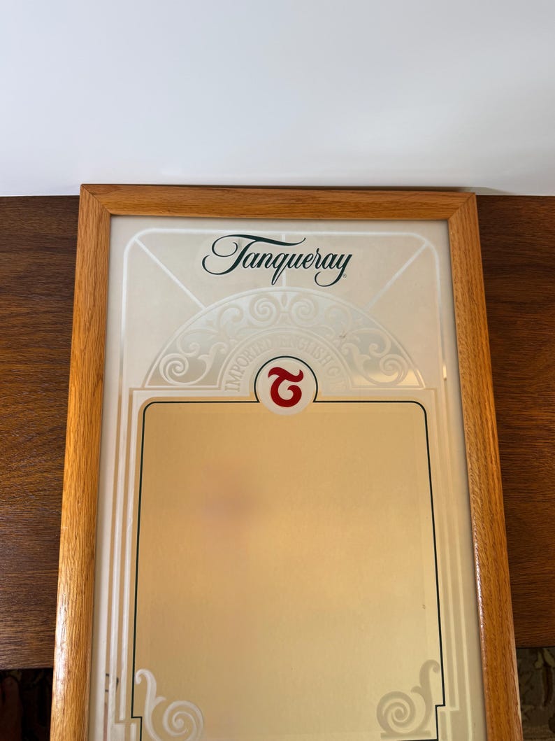 May include: A framed Tanqueray gin advertisement. The wooden frame surrounds a cream-colored background with the Tanqueray logo in elegant script. The bottom section is a gold color. The words "IMPORTED ENGLISH GIN" are visible.