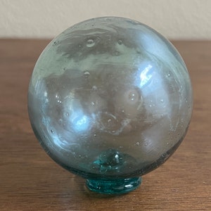 May include: A round, light blue glass fishing float with a small, round base. The float has many small air bubbles trapped inside.