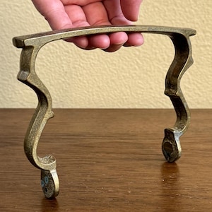 May include: A brass handle with a unique, ornate design. The handle has a curved shape and two decorative ends.