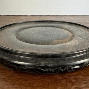 May include: A round, black wooden pedestal with a carved design around the edge. The pedestal is made of dark wood and has a smooth, polished surface.