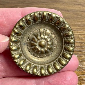 May include: A circular brass button featuring a central raised floral motif surrounded by a ring of textured, oval-shaped indentations. The metal surface displays a weathered, antique gold finish with darkened patina in the recessed areas.