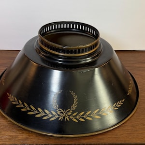 Könnte beinhalten: Ein schwarzer Metall-Lampenschirm mit einem goldfarbenen Lorbeerblatt-Design am Sockel. Der Lampenschirm hat eine ausgestellte Form und eine kreisförmige Oberseite mit einer Reihe von vertikalen Schlitzen. Der Rand ist ebenfalls goldfarben.