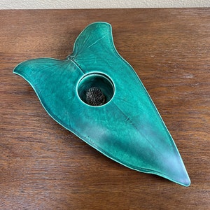 May include: A green ceramic leaf-shaped vase with a round opening in the center. The vase is designed to hold a single flower or small bouquet.