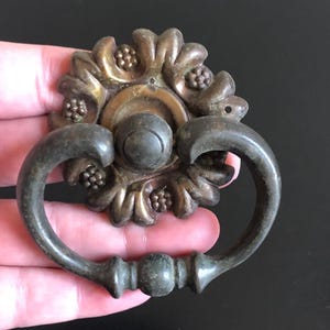 May include: Antique bronze drawer pull with a floral design. The pull features a circular backplate with a sunburst pattern and a curved handle. The hardware is made of metal and has a dark, aged finish.