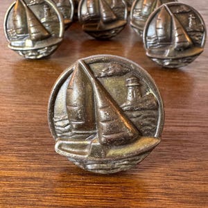 May include: Bronze-toned cabinet knobs with a nautical theme. Each knob features a sailboat and lighthouse design. The knobs are round and have a detailed, antique appearance, perfect for coastal decor.