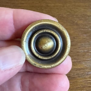 May include: A round, brass cabinet knob with a concentric circle design. The knob has a slightly aged, antique finish.
