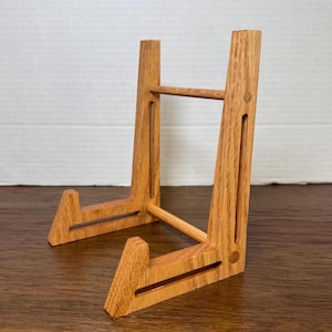 Könnte beinhalten: Ein hölzerner Präsentationsständer mit natürlichem Finish. Der Ständer hat ein A-Rahmen-Design mit einer horizontalen Stützstange und abgewinkelten vorderen Stützen. Der Ständer ist für die Aufnahme einer Platte oder eines anderen dekorativen Gegenstands konzipiert. Die Holzmaserung ist sichtbar.